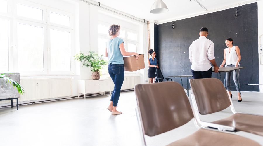 One person moving a box, two people moving a table, and one person moving a chair into an office. These jobs are included on a 'how to move an office' checklist. 